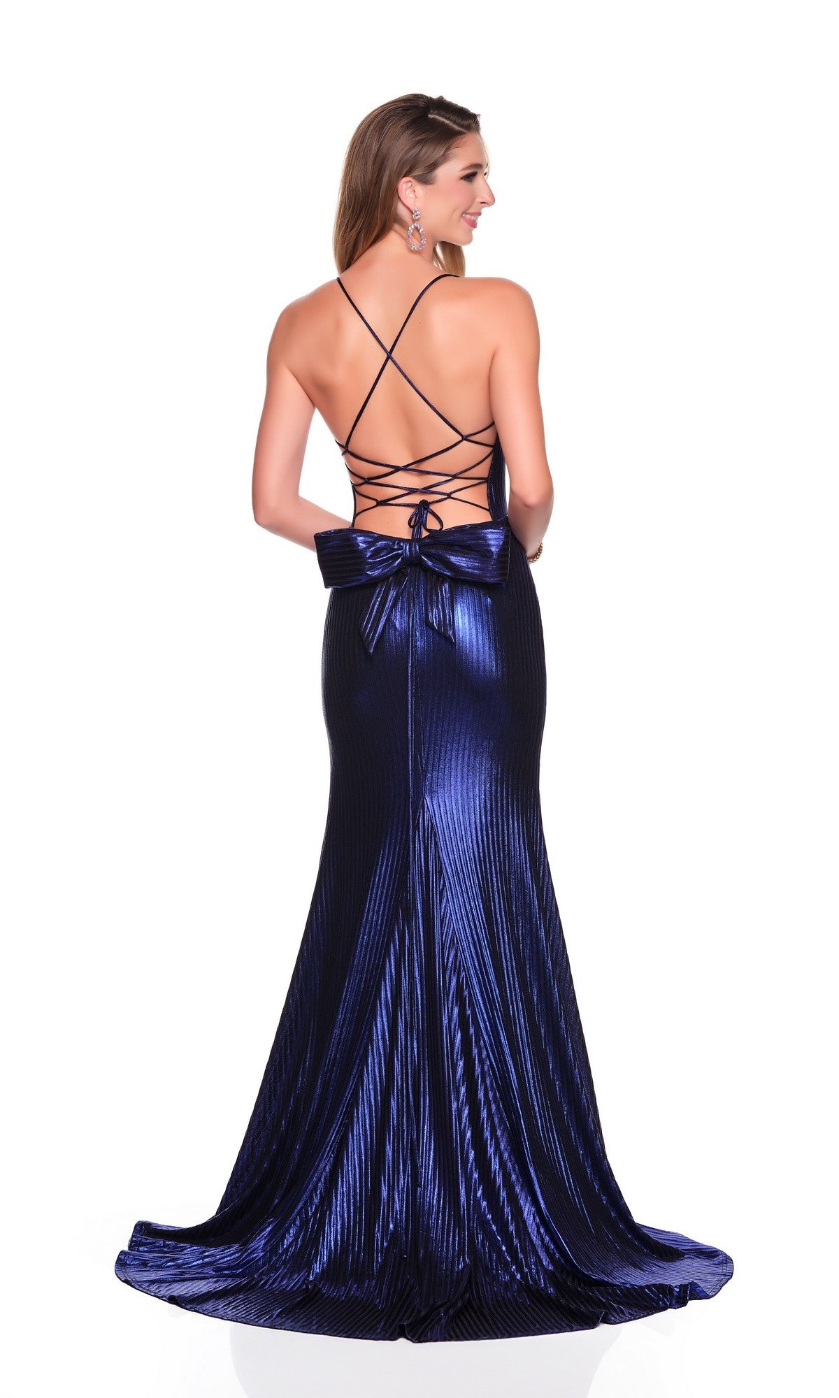 A woman wears the Dave and Johnny 12734 long prom dressโa floor-length, dark blue metallic gown featuring a corset bodice, strappy open back, and large bow at the lower backโstanding against a white background.